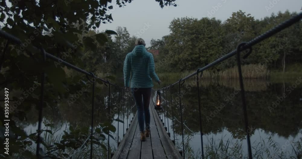 Woman walking in nature over river on suspension bridge in holding old lantern Woman walking in nature over river on suspension bridge in holding old lantern