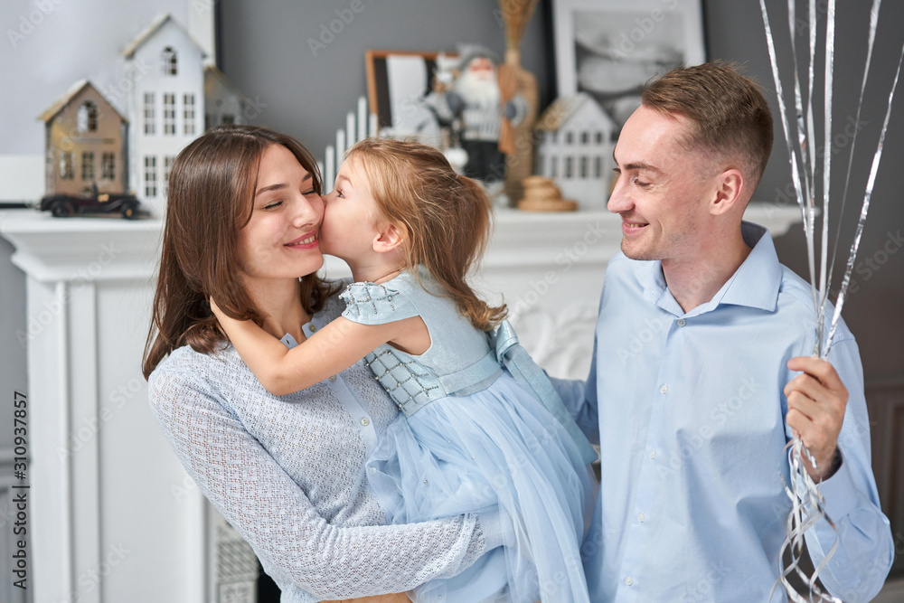 Fototapeta premium Loving family. Mom and dad hugging little daughter . Parents and baby child having fun near Christmas tree and white fireplace indoors. Merry Christmas and Happy New Year. Cheerful pretty people.