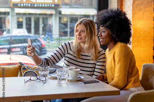 Two multi ethnic girl friends enjoying coffee together in a coffee shop, sitting at a table and looking pictures or social media content on a smartphone.