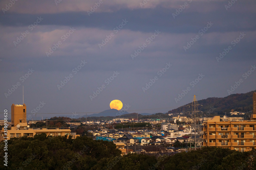 Fototapeta premium Full moon rises over distant mountain in small Japanese town at dusk