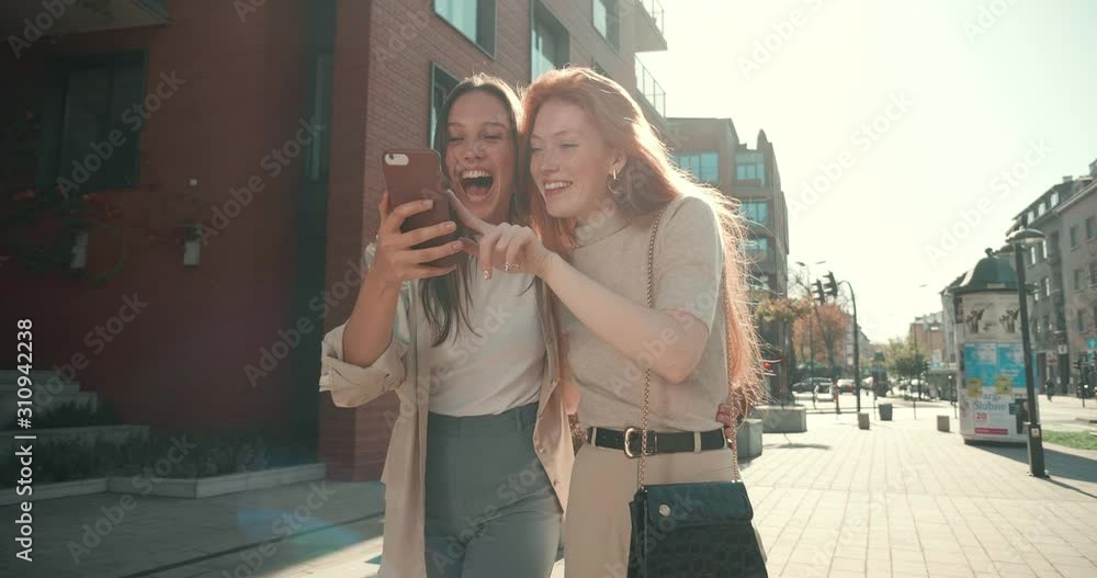 Couple of Female Young Friends walking in the Town while looking at Mobile Phone. Pretty women having Fun in the Center City. Smiling and Laughing while hugging each other. Happy Frienship. Couple of Female Young Friends walking in the Town while looking at Mobile Phone. Pretty women having Fun in the Center City. Smiling and Laughing while hugging each other. Happy Frienship.