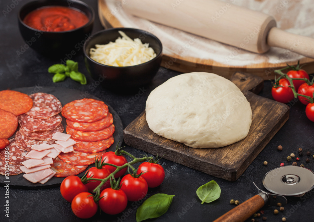 Preparation for baking of pepperoni pizza with raw dough, salami spicy chorizo with wheel cutter and fresh tomatoes and basil on dark background with bowl plates with cheese and tomato paste. Preparation for baking of pepperoni pizza with raw dough, salami spicy chorizo with wheel cutter and fresh tomatoes and basil on dark background with bowl plates with cheese and tomato paste.