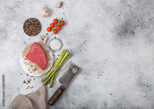 Slice of Raw Beef sirlion steak on round chopping board with tomatoes,garlic and asparagus tips and meat hatchet. Space for text photo