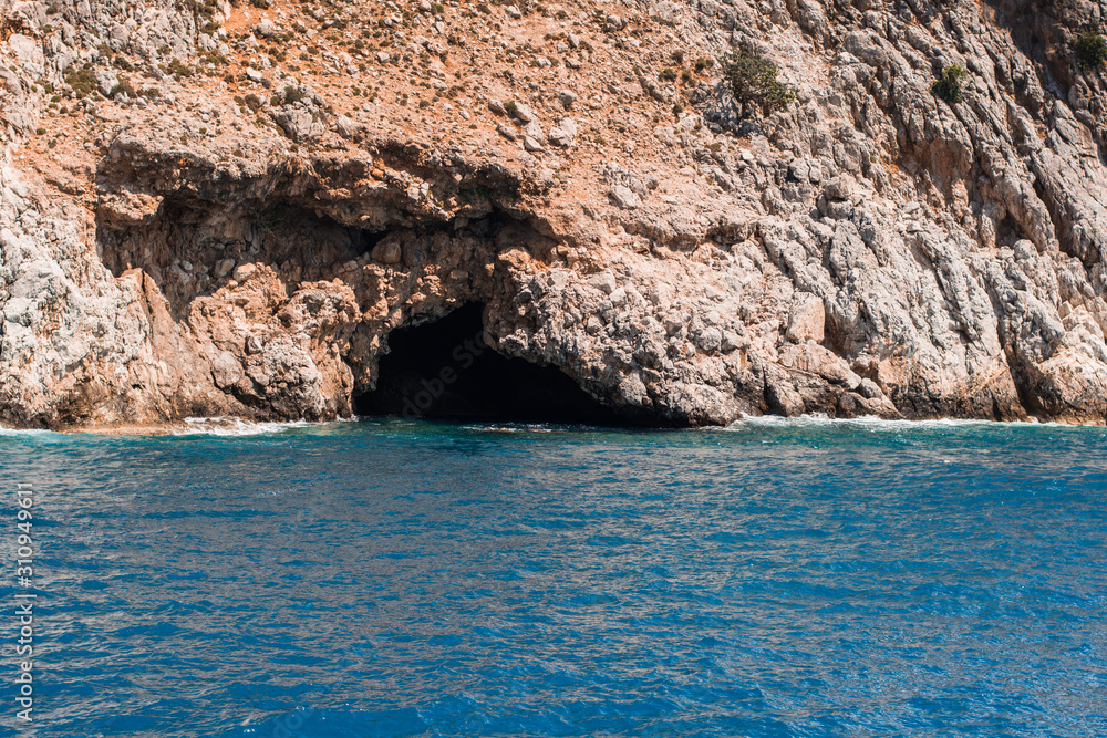 Photo of a cave, looking dark and cold, in a rocky sea coast on sunny summer day.