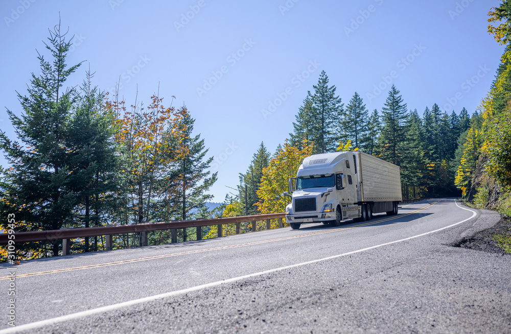 Big rig white bonnet semi truck with refrigerator semi trailer driving down hill on the mountain autumn road with trees on the hillside