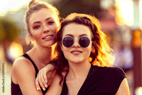 Close up portrait of young female couple. Two attractive girls looking at camera while relaxing outdoors, admiring sunset. Lesbian lives, summertime concept. Horizontal shot. Selective focus