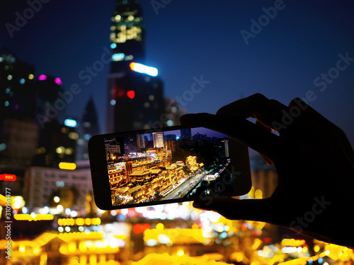 Silhouette hand holding mobile phone taking photograph night landscape of Hongyadong shopping complex at Chongqing, China photo