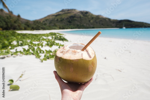 Woman's hand holding fresh coconut with bamboo straw on a beautiful white sandy beach in Fiji with turquoise water, green hills and plants, relaxing vacation destination