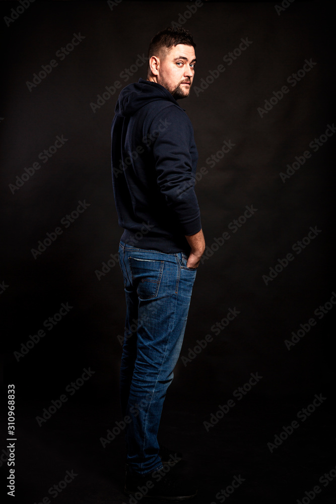 Handsome young man in jeans with a beard. Black background. Vertical.