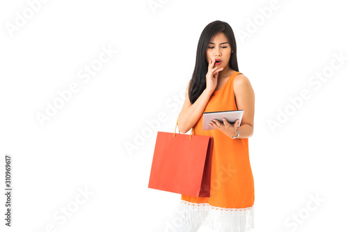 A beautiful Asian young woman wearing orange dress holding a tablet and a shopping bag looking frustrated or confuse in front of white isolated background.