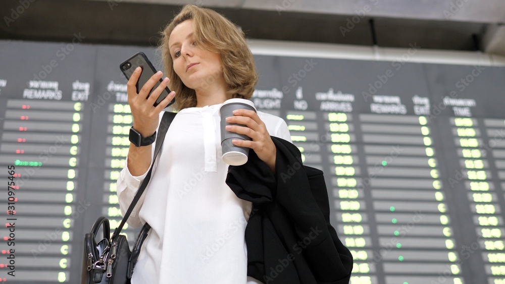 Fototapeta premium Young Businesswoman Using Smartphone At Departure Timetable Display At Airport