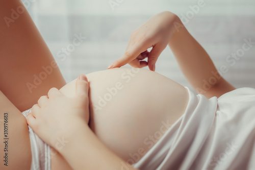 A beautiful pregnant woman in white underwear is lying on her bed and gently stroking her belly. Great time to wait for the baby. Close-up photo of a pregnant belly