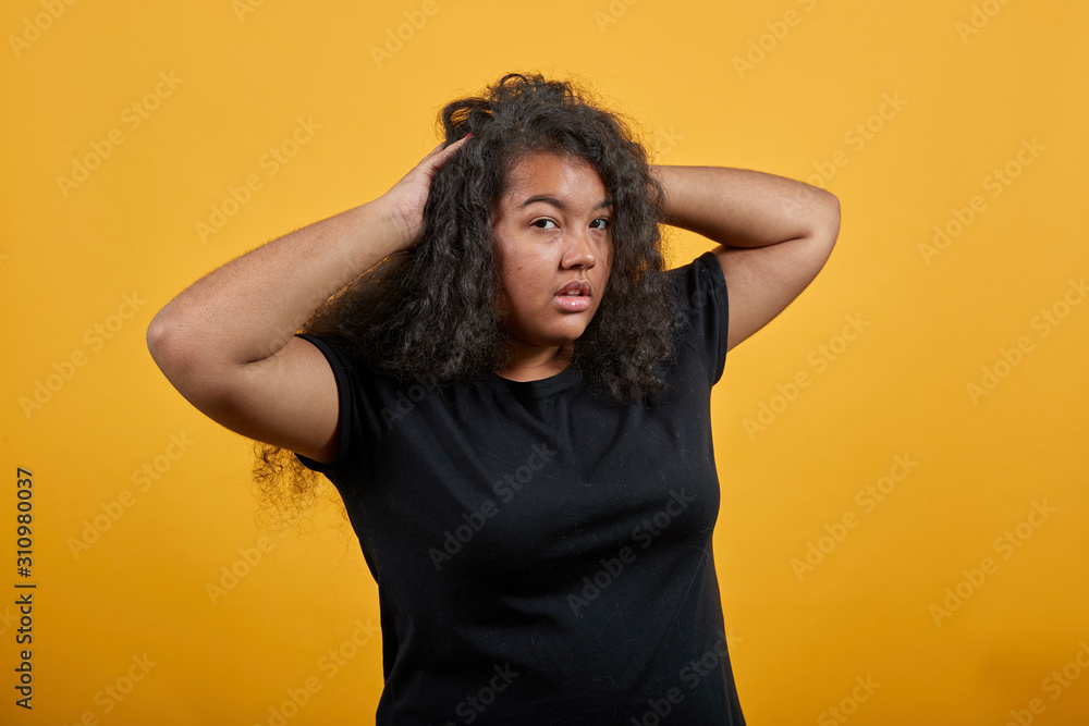 Confused afro-american young woman with overweight over isolated orange background wearing fashion black shirt keeping hands on head, corrects haircut. People lifestyle concept. Confused afro-american young woman with overweight over isolated orange background wearing fashion black shirt keeping hands on head, corrects haircut. People lifestyle concept.
