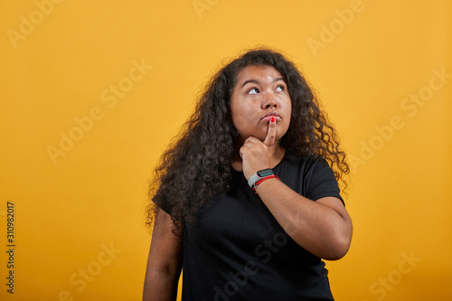 Strict afro-american young woman with overweight keeping finger on mouth, looking up, thinking, over isolated orange background wearing fashion black shirt. People lifestyle concept.
