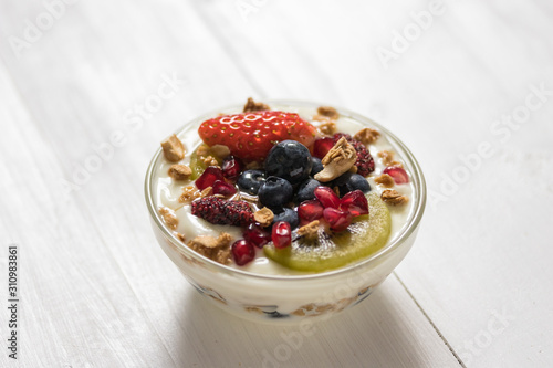 Plain yogurt with strawberry, blueberries, kiwi, granola, pomegranate in a glass bowl and honey on white wooden texture, healthy food and plant-based food concept
