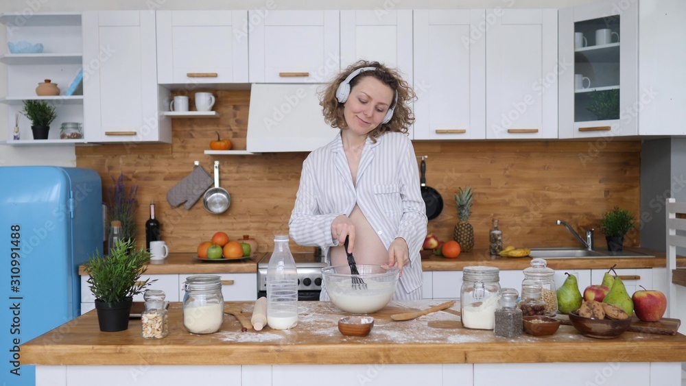Happy Healthy Pregnancy Concept. Pregnant Woman In Headphones Dancing And Cooking On Kitchen.
