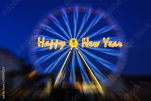 Happy New Year sparkling text over a spinning ferris wheel in motion blur against a dark blue sky, copy space