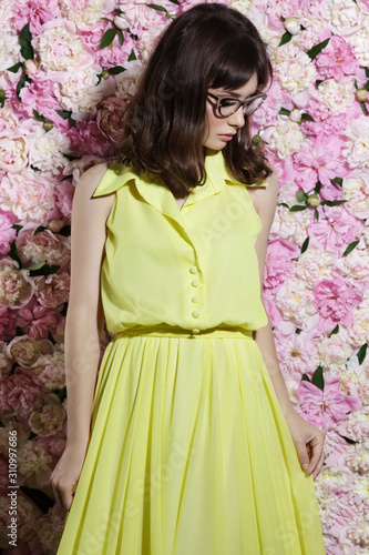 Young woman at the peonies wall, wear in yellow long dress. Fashion, make-up content.