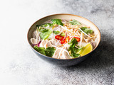 Traditional Vietnamese soup- pho ga in bowl with chicken and rice noodles, mint and cilantro, red onion, chili, bean sprouts and lime on grey background. Asian food. Copy space.