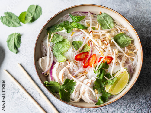 Traditional Vietnamese soup- pho ga in bowl with chicken and rice noodles, mint and cilantro, red onion, chili, bean sprouts and lime on grey background. Asian food. Top view