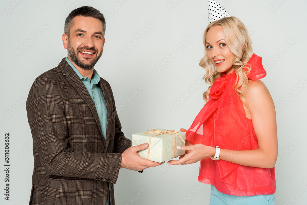 smiling man and happy wife smiling at camera while holding gift box isolated on grey smiling man and happy wife smiling at camera while holding gift box isolated on grey