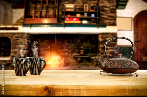 Metal kettle on wooden table.Free space for your decoration.Blurred background of fireplace with orange fire. Cold december night in home interior.Hot drink of tea,coffee or mulled.