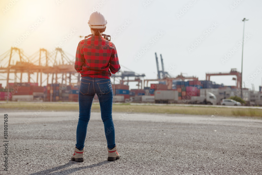 Back view, Young female engineer in protective work white wear in shipping yard examining cargo, background cargo, transport cargo engineer worker concept.