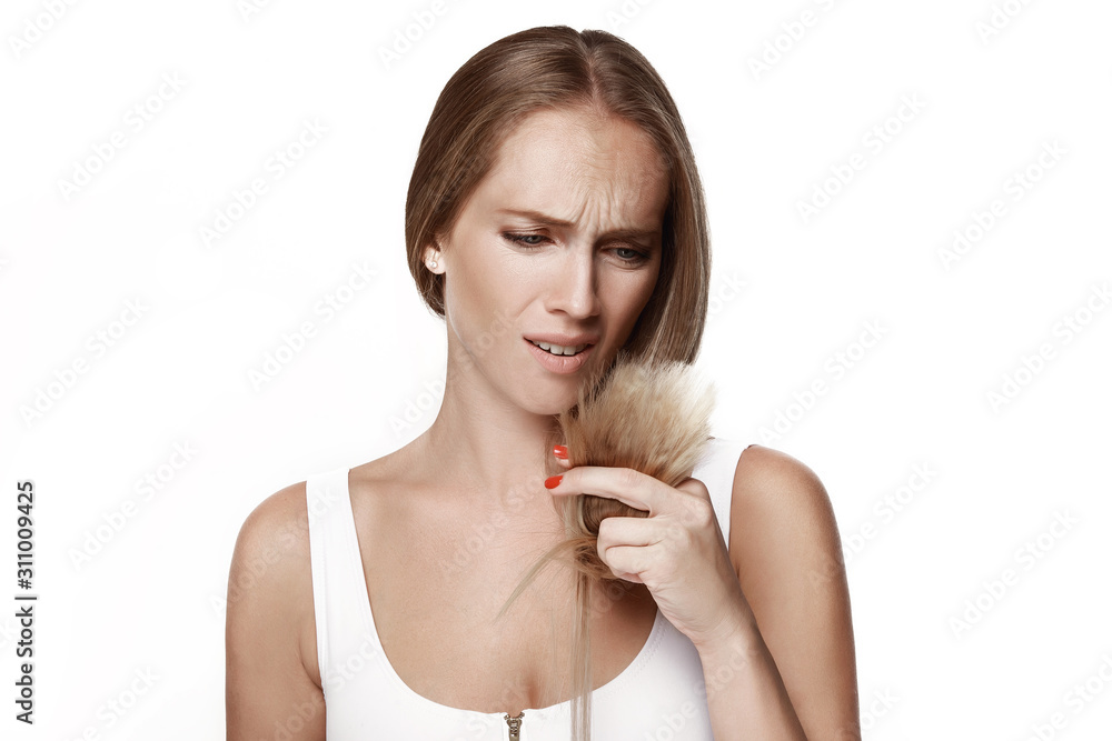 A beautiful girl looks at her hair, holding it in her hands. Emotions, disappointment, surprise. Portrait on a white background. A beautiful girl looks at her hair, holding it in her hands. Emotions, disappointment, surprise. Portrait on a white background.
