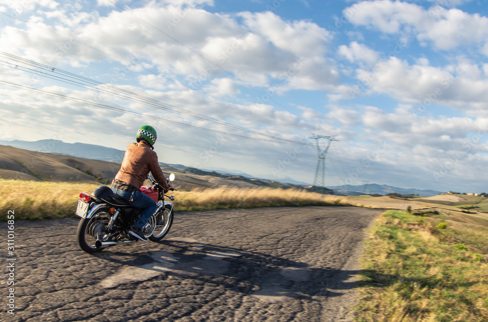 Fototapeta premium Man riding motorbike along a country road on tuscan hills landscape at sunset. Tuscany, Italy.