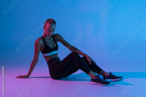 Full length portrait of attractive woman wearing black tracksuit doing gymnastic sitting on the floor isolated over purple background