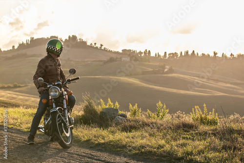 Man on his motorbike standing on a country road on hills landscape at sunset. Tuscany, Italy. photo