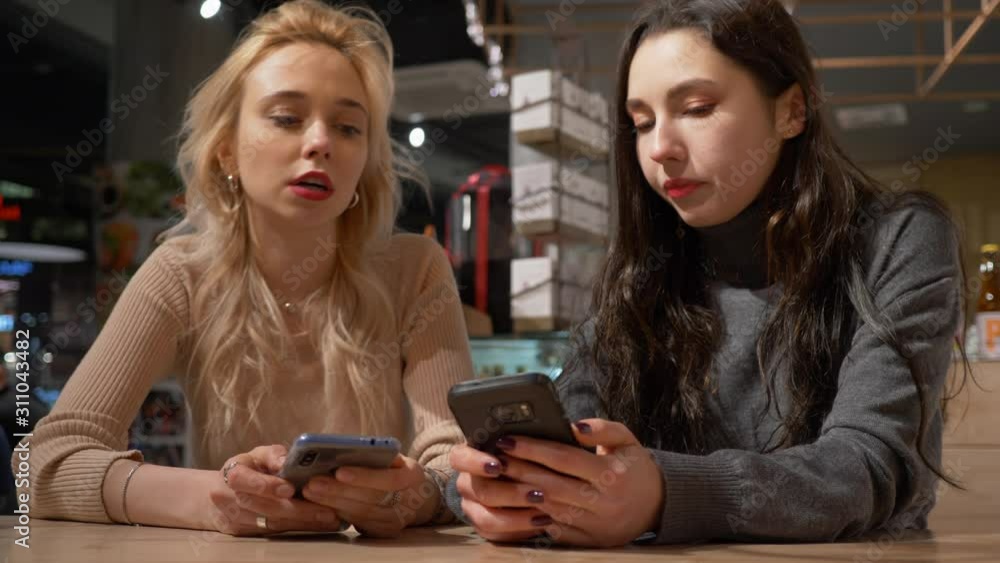 Two young pretty women girlfriends watch use mobile phones smartphones surfing internet social media while sitting in cafe waiting an order Two young pretty women girlfriends watch use mobile phones smartphones surfing internet social media while sitting in cafe waiting an order