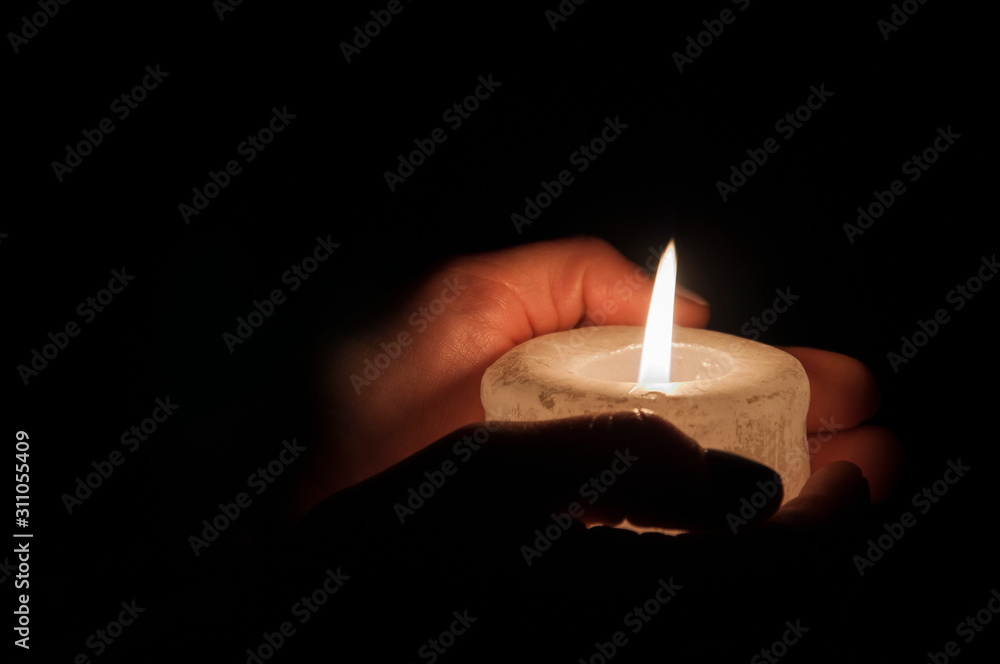 A large candle burns in the hands in the dark. On a wooden background. A large candle burns in the hands in the dark. On a wooden background.