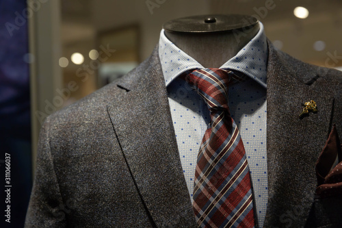 A dark gray jacket, a tie with blue and Burgundy stripes, a blue shirt with a pattern of squares hang on a mannequin behind glass in the window of a fashionable men's clothing store.