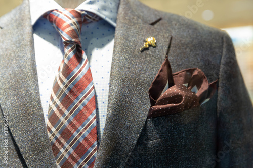 A dark gray jacket, a tie with blue and Burgundy stripes, a blue shirt with a pattern of squares hang on a mannequin behind glass in the window of a fashionable men's clothing store.