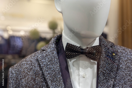 A gray coat, a blue jacket, a dark shade and a white shirt with a bow tie with a square pattern hang on a mannequin behind glass in the window of a fashionable men's clothing store.