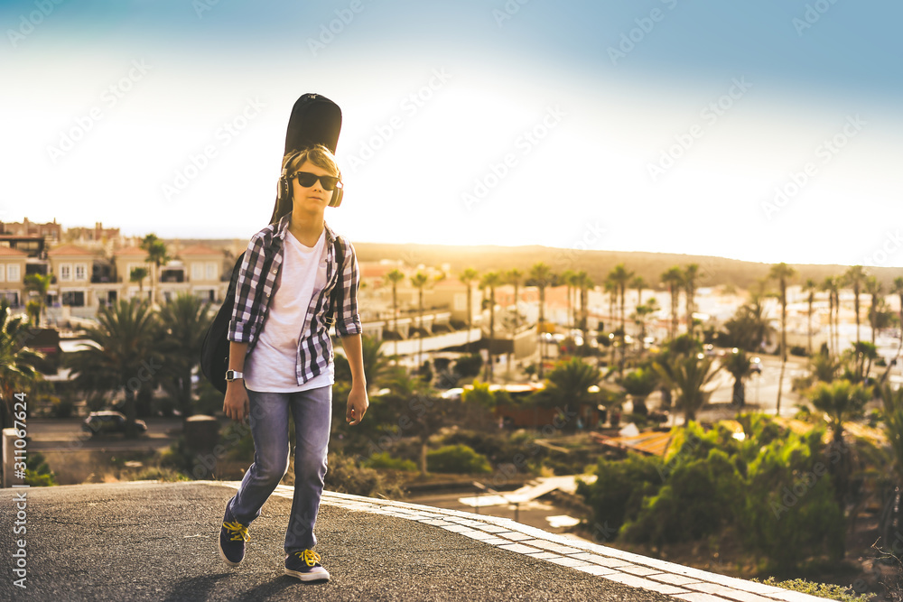 Fototapeta premium Trendy young boy walking outdoor with guitar on his shoulders like a backpack. Teen listening to music come back from school, exotic panorama and sunset in background. Freedom, education youth concept