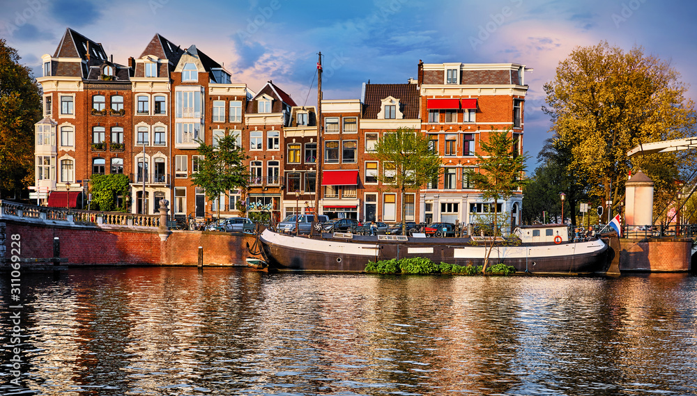 Amsterdam, Netherlands. Floating House and houseboat at channels by banks. Traditional dutch dancing houses among trees. Evening autumn street above water pink sunset sky with clouds.