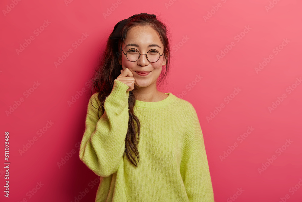 Pretty cheerful girl with dark hair, keeps hand on cheek, has pleased face expression, dressed in green jumper and round spectacles, smiles pleasantly, isolated on pink background, expresses happiness Pretty cheerful girl with dark hair, keeps hand on cheek, has pleased face expression, dressed in green jumper and round spectacles, smiles pleasantly, isolated on pink background, expresses happiness
