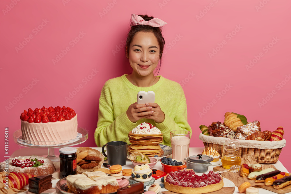 Calories, appetite and dieting concept. Satisfied Asian girl has tasty snack, sugar addiction, assortment of unhealthy products on table bad for figure and teeth, enjoys treat, uses cellular