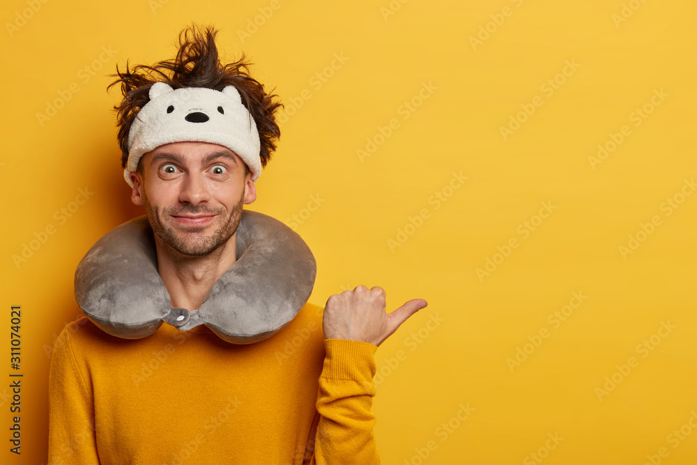 Glad European man points thumb away, dressed in yellow casual jumper, demonstrates blank space, wears travel pillow, travels in plane. People, traveling lifestyle, departure, voyage concept.