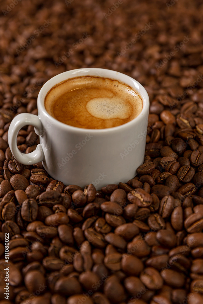 A small white cup of coffee stands on coffee beans. Fragrant pleasure. Background. Space for text. Vertical. A small white cup of coffee stands on coffee beans. Fragrant pleasure. Background. Space for text. Vertical.