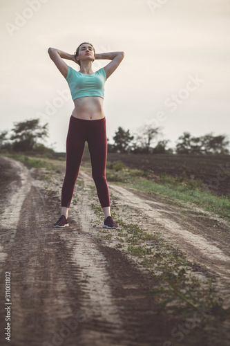 slim young woman in sportswear exercises and stretches hands up in the field, girl engaged in sport outdoors on a cloudy day, concept healthy lifestyle and female beauty