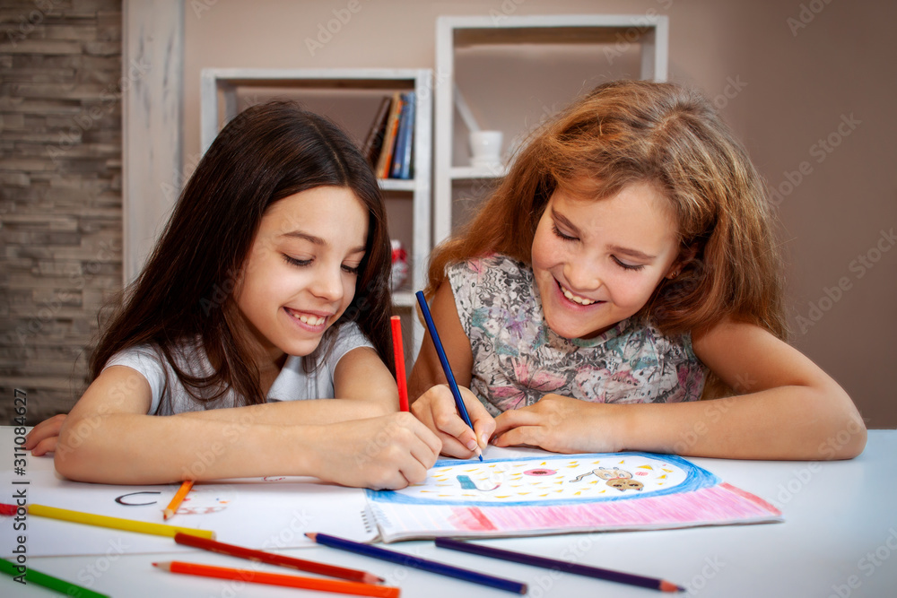two beautiful teenage girls, a brunette and a blonde laugh, draw with colored pencils in an album on a white table, in the background are white shelves with books and toys two beautiful teenage girls, a brunette and a blonde laugh, draw with colored pencils in an album on a white table, in the background are white shelves with books and toys