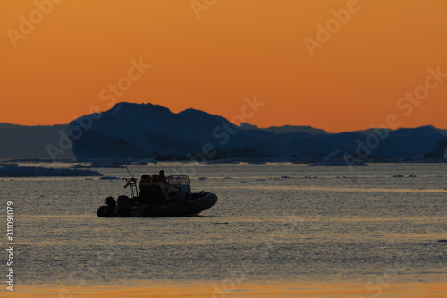 Silhouetted inflatable boat with passengers on calm Arctic waters at sunset, icebergs in the background. photo