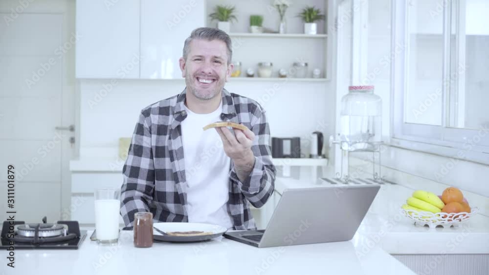 Funny Caucasian man laughing in the kitchen while holding a bread with laptop on the table. Shot in 4k resolution Funny Caucasian man laughing in the kitchen while holding a bread with laptop on the table. Shot in 4k resolution