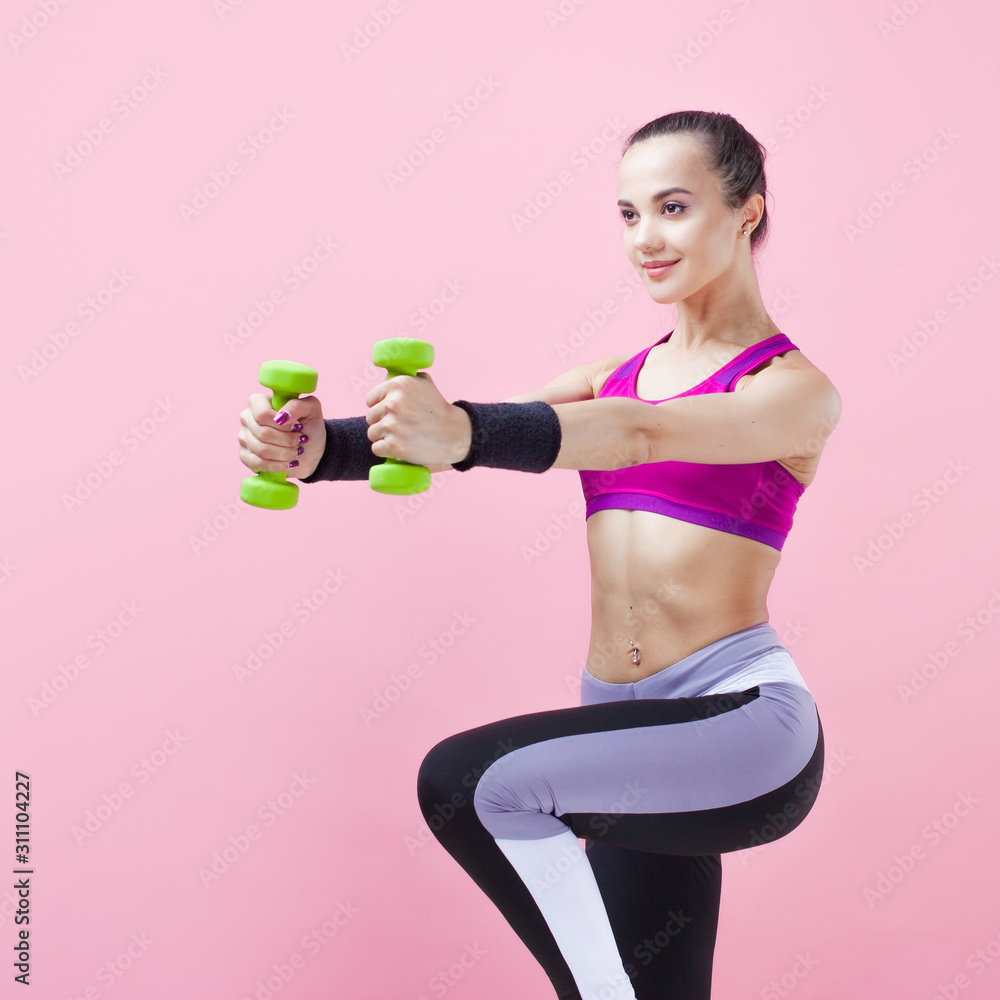 Young athletic woman, in a bright sports top, with green dumbbells, raises her arms and knees, on a pink background. Young athletic woman, in a bright sports top, with green dumbbells, raises her arms and knees, on a pink background.