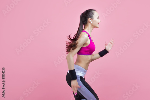 A young attractive girl runs with smile, wireless headphones and in a bright sports top, on a pink background, cardio. © Ulia Koltyrina