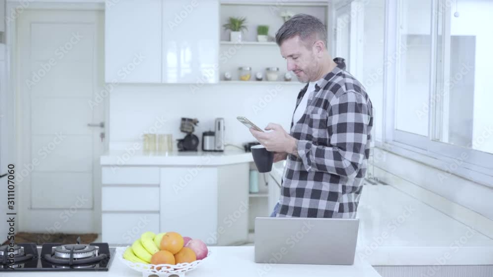 Caucasian man standing in the kitchen while using a mobile phone and drinking a cup of coffee. Shot in 4k resolution Caucasian man standing in the kitchen while using a mobile phone and drinking a cup of coffee. Shot in 4k resolution