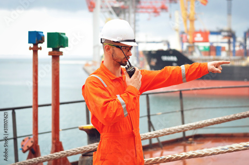 An experienced sailor in orange overalls and a white helmet speaks on a walkie-talkie and gives instructions to other workers.